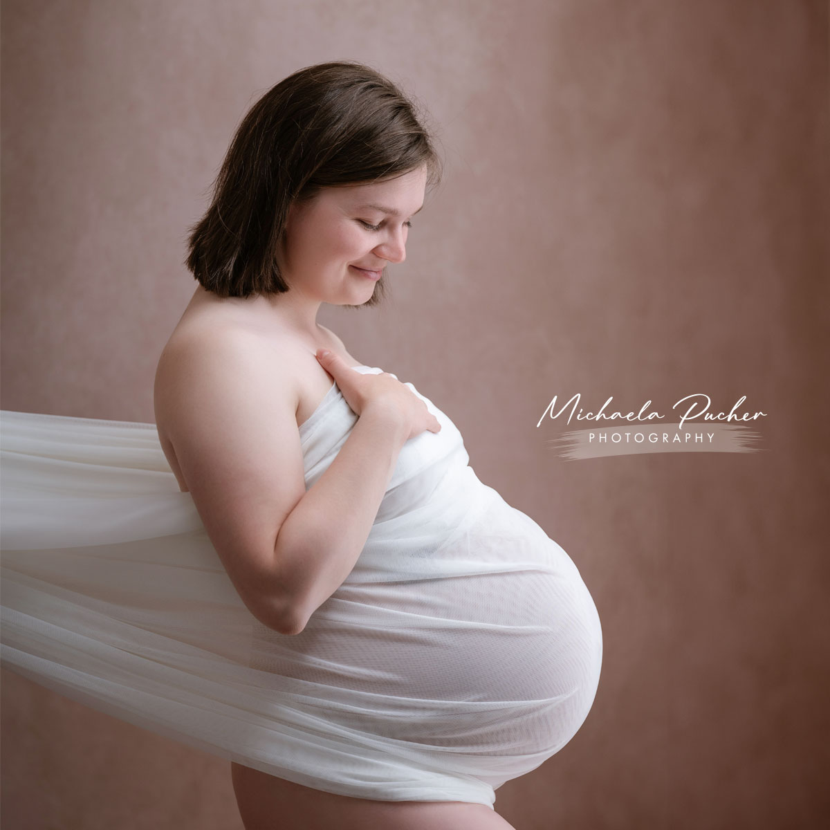 Hochschwangere Frau im Studio, seitlich fotografiert, eingehüllt in einen weißen, transparenten Stoff, lächelt sanft und hält eine Hand auf ihrem Babybauch.
